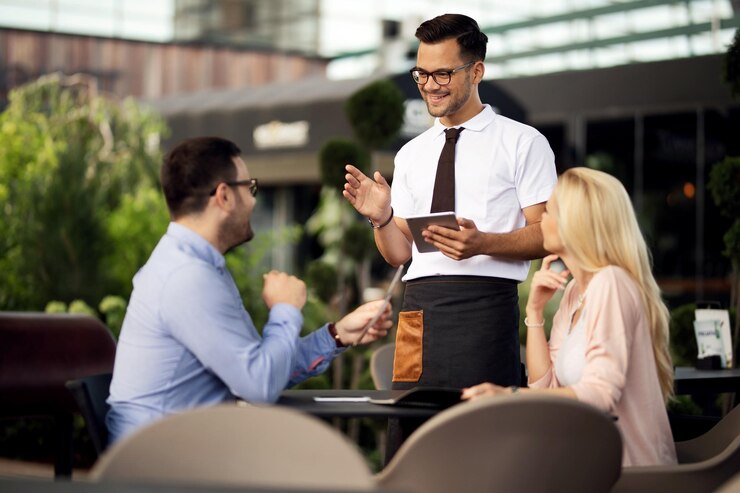 young happy waiter using digital tablet while talking couple taking their order cafe
